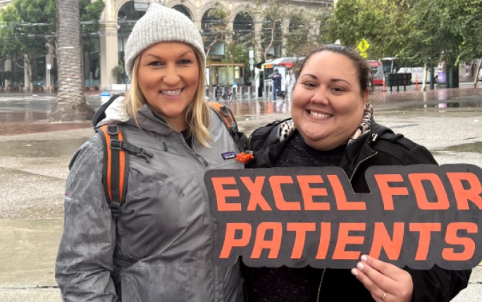 Two female Exelixis employees smiling, one holding an “Excel for Patients” sign