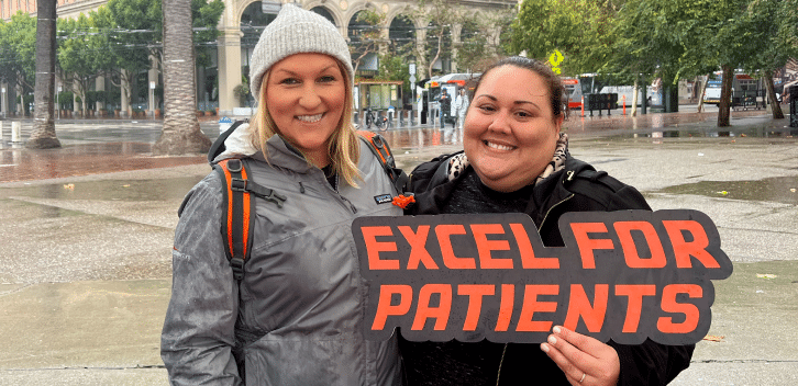 Two female Exelixis employees smiling, one holding an “Excel for Patients” sign